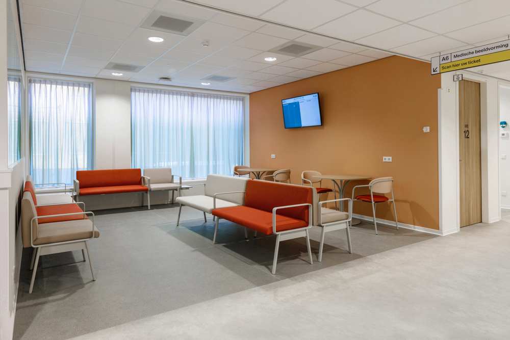 Moderne wachtruimte in ziekenhuis SJG Weert met zitbanken, stoelen, tafels, informatiescherm en bewegwijzering.