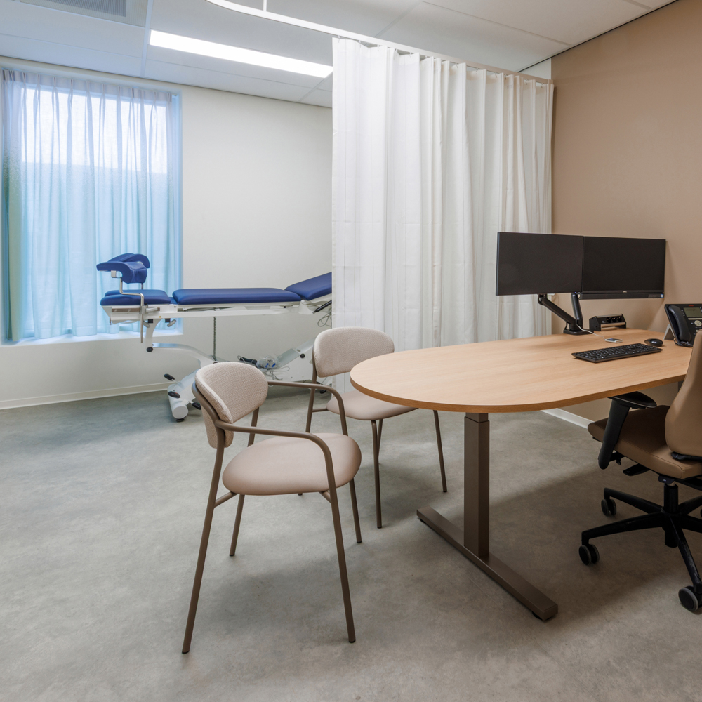 Moderne onderzoekskamer in ziekenhuis SJG Weert met behandelbank, bezoekersstoelen, bureau, computerschermen en privacygordijn.