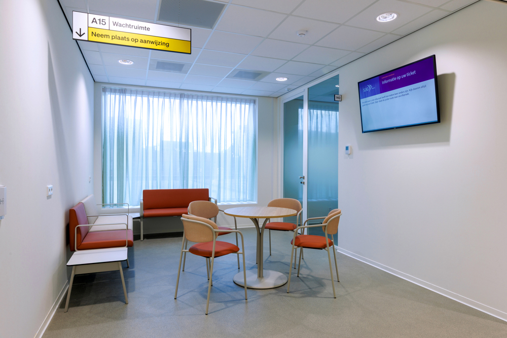 Wachtruimte in een zorgomgeving met stoelen, bank, ronde tafel en digitale informatiescherm.